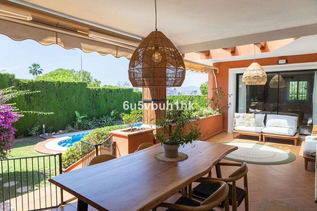Outdoor dining patio with a pool and garden view. Features a rustic wooden table and hanging rattan lights.