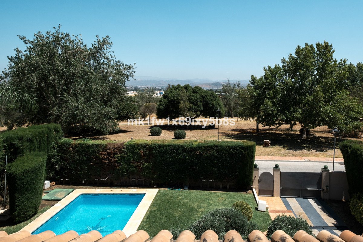 A clear blue swimming pool and lush green garden with trees and a distant view of hills under a bright blue sky.
