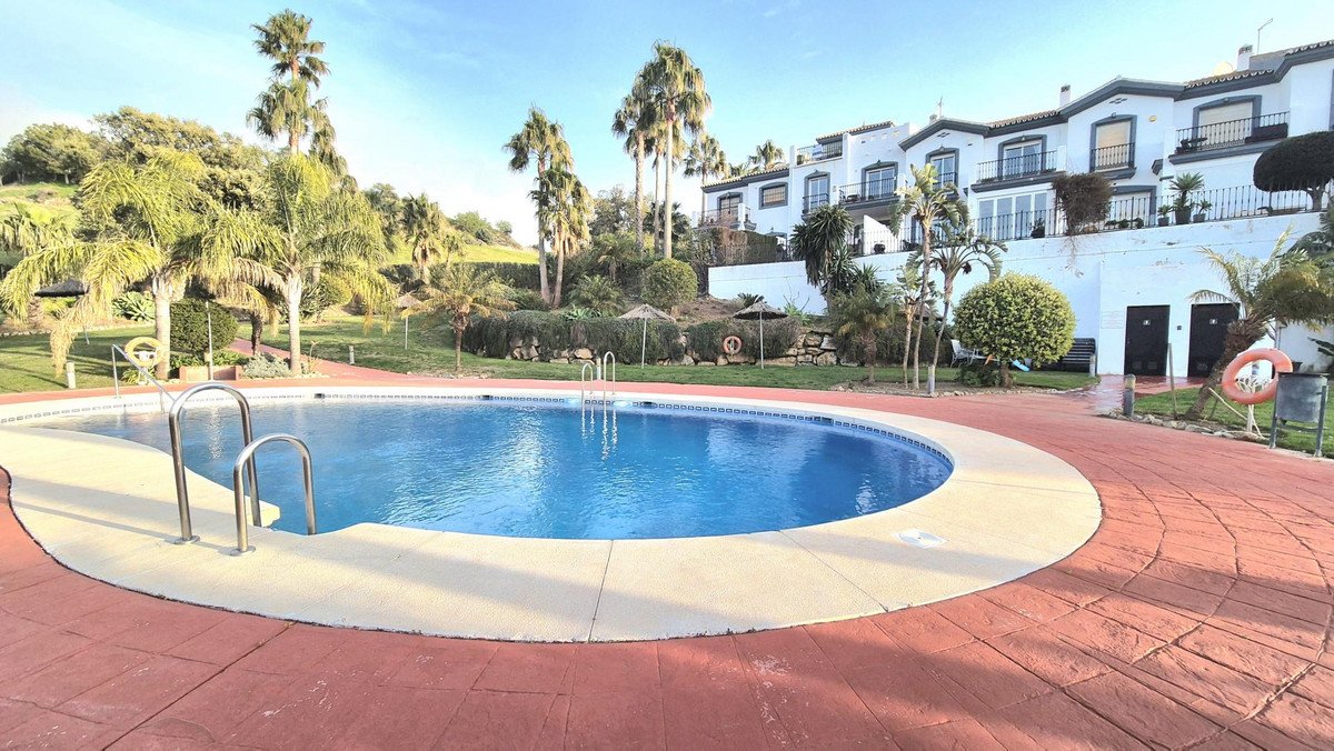 A bright outdoor swimming pool with a complex of white buildings and palm trees in the background under a clear blue sky.