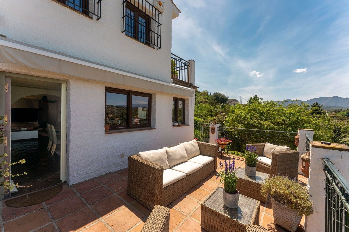 Terrace with wicker outdoor seating furniture, coffee tables, and a view of lush green trees and distant mountains under a blue sky.