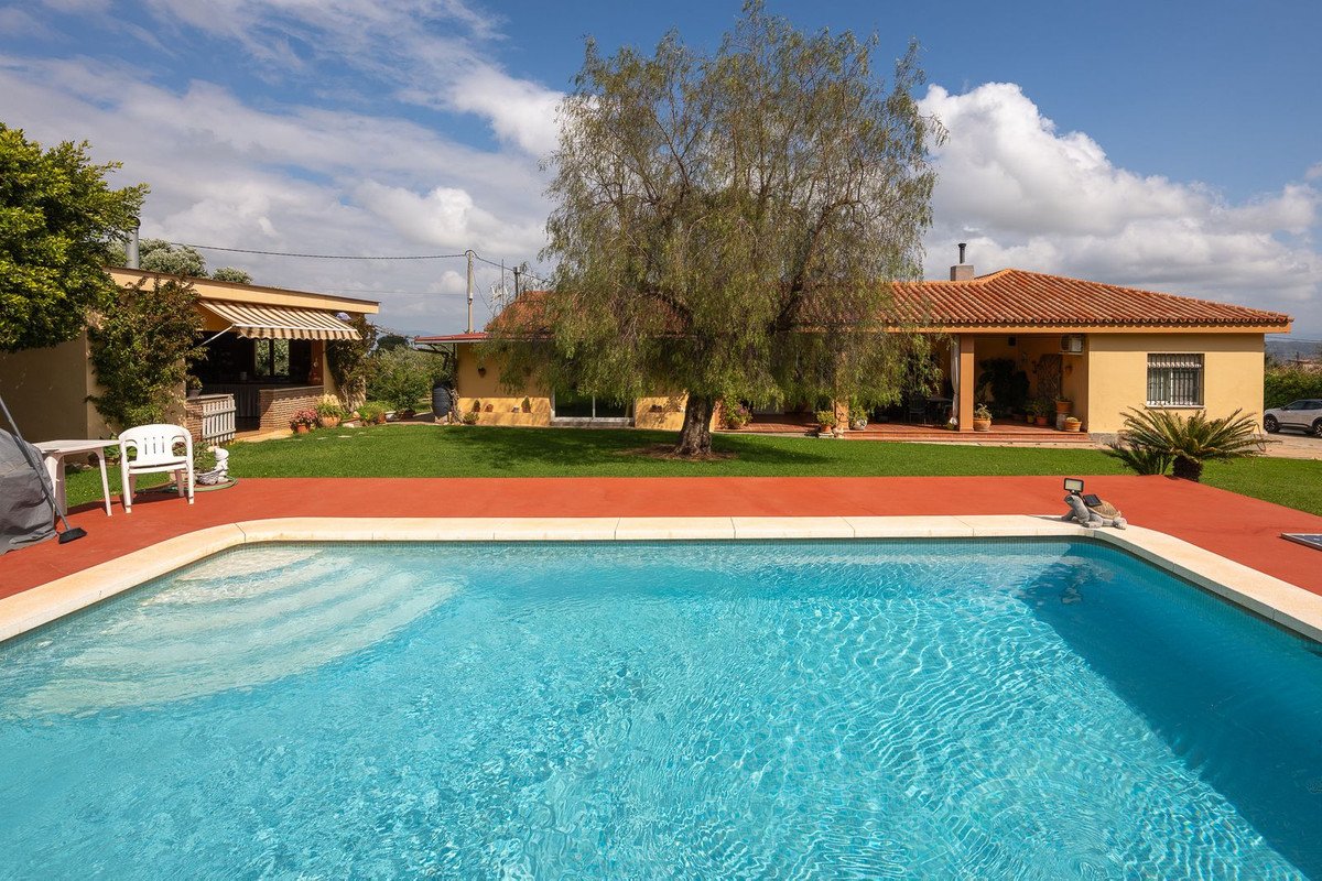 A sparkling blue swimming pool in the foreground, with a yellow villa and lush green garden under a partly cloudy sky.