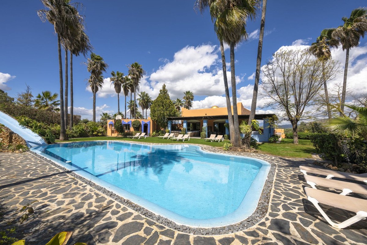 Villa with pool, palm trees, and lounge chairs under blue sky.