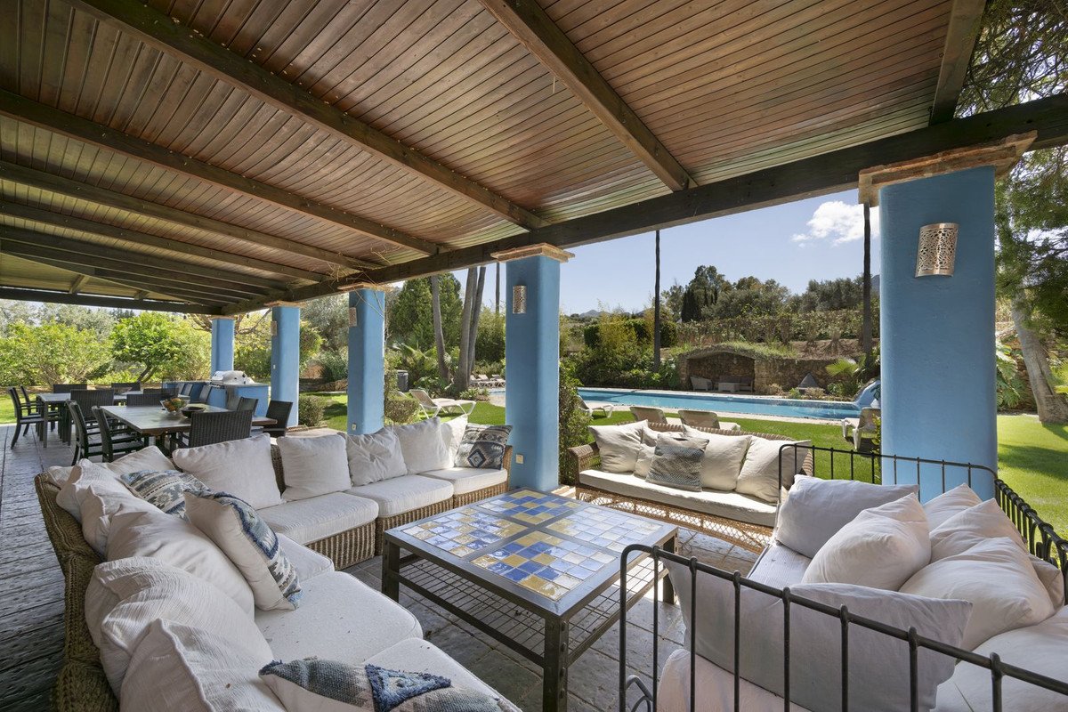 Covered patio with wicker sofas, blue pillars, and a view of a pool and green garden.