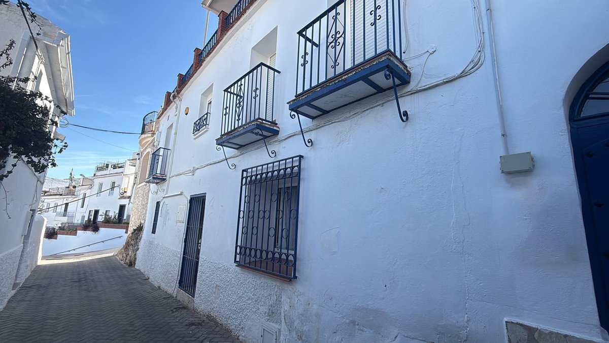 A narrow street in a town with white buildings. The building on the right has multiple balconies with dark railings.