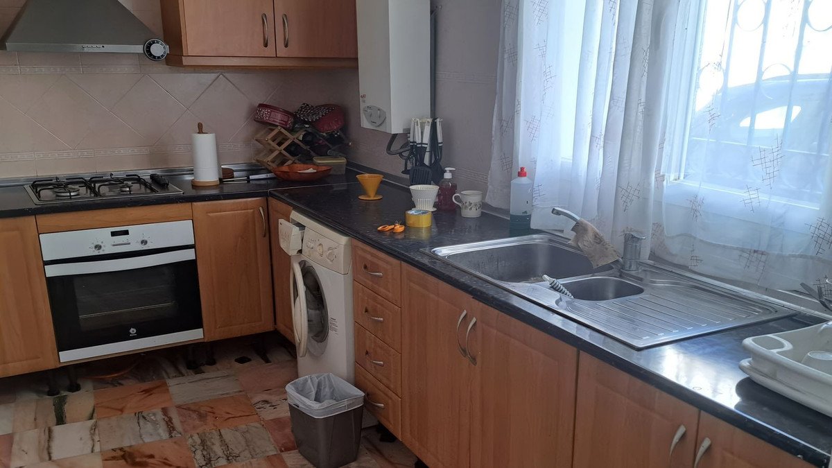 A well-lit kitchen featuring a gas hob, oven, sink, washing machine, and wooden cabinets under a window.