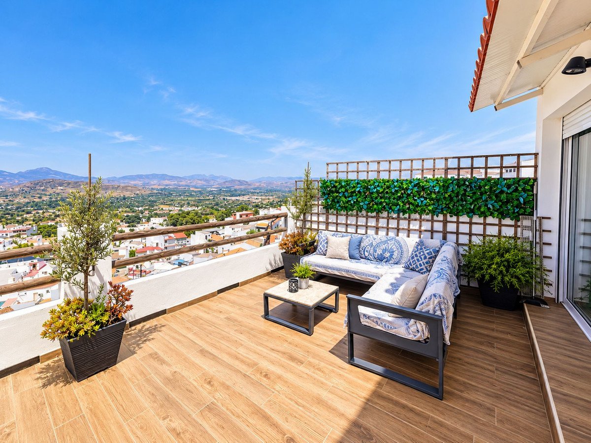 A spacious terrace with outdoor seating, coffee table, and plants offering panoramic views of a town and mountains under a clear blue sky.