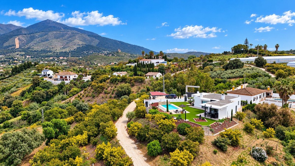 Modern villa with swimming pool and green lawn set in a hilly landscape with mountains and trees under a blue sky.