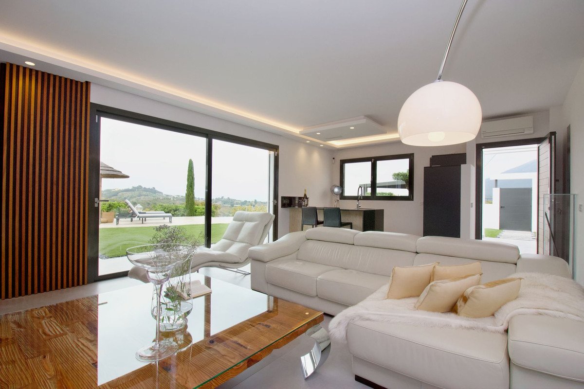 Modern living room with a large white sectional sofa, glass coffee table, and sliding glass doors offering a view of a pool and garden.