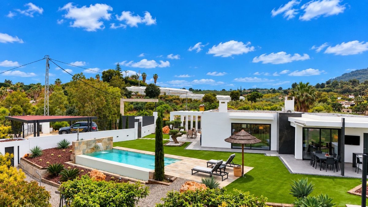 Modern villa with swimming pool, outdoor dining area, and lush green garden under a blue sky with white clouds.