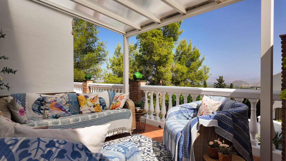 A covered balcony with comfortable seating, colourful cushions, and a scenic mountain view under a clear blue sky.
