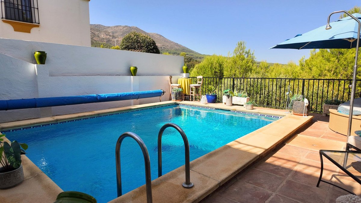 A swimming pool with a blue cover sits beside a patio with terracotta tiles and a view of green hills and mountains.