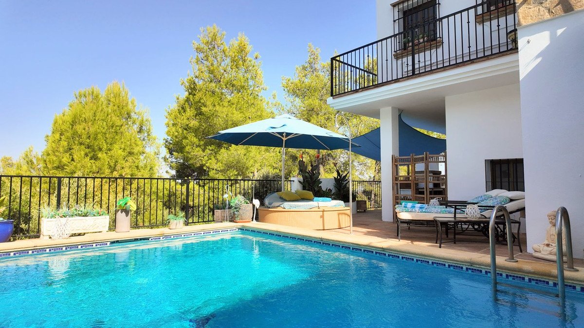 Outdoor swimming pool with lounge chairs, a daybed, and a large umbrella on a sunny day with trees and a balcony visible.