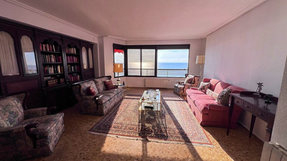 Spacious living room with large windows overlooking the sea, featuring a bookshelf, sofas, and armchairs.