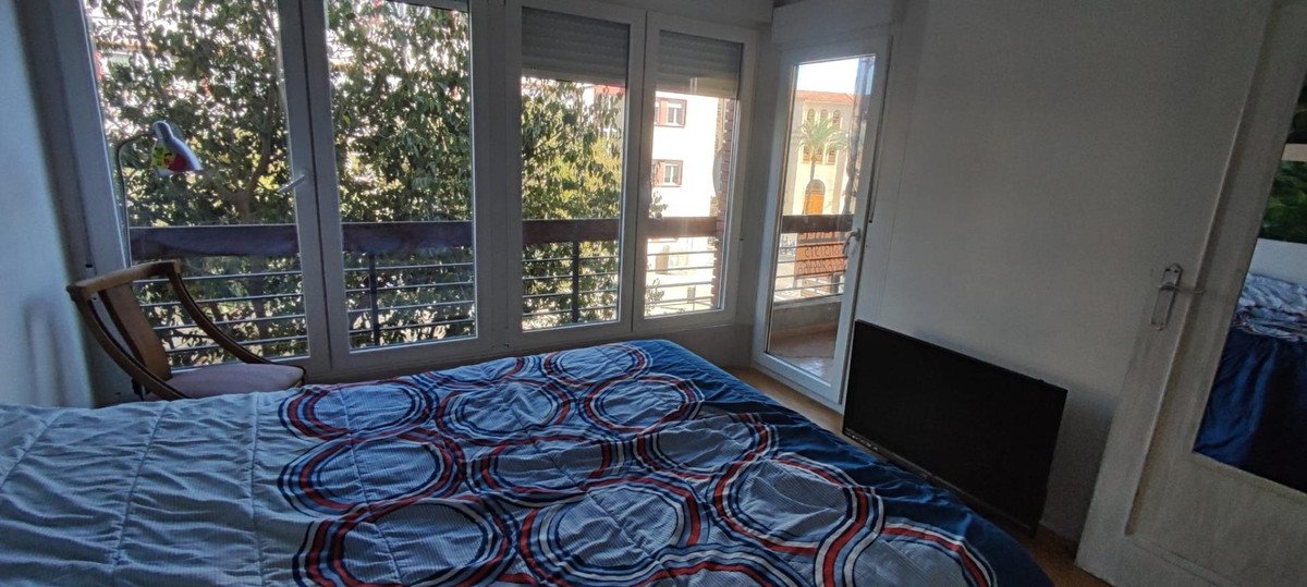 Bedroom with patterned bedspread, large windows, balcony view of trees and buildings.