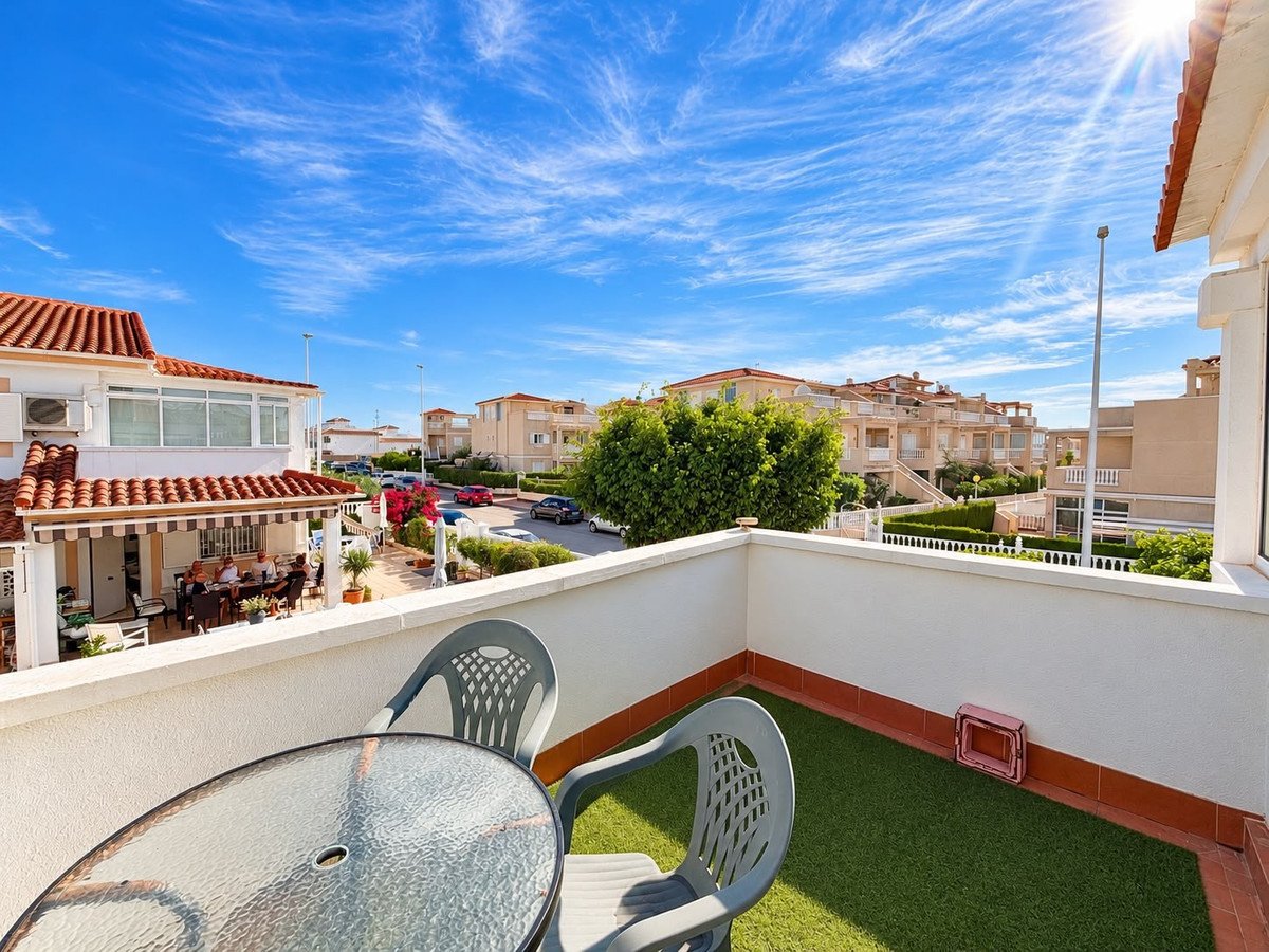 A terrace with a table and chairs offers a view of houses and a street under a blue sky with clouds.