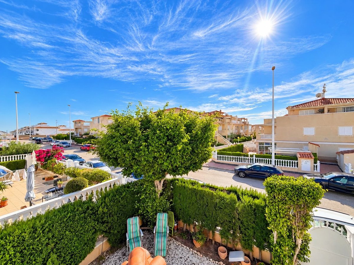 Terrace with chairs overlooking a street lined with houses under a bright, sunny sky with wispy clouds.