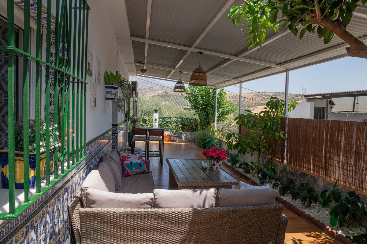 A covered patio with wicker seating, a coffee table with flowers, and a view of rolling hills and olive trees.