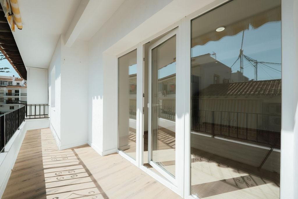 A bright balcony with light wood flooring and large sliding glass doors offering a view of residential buildings.
