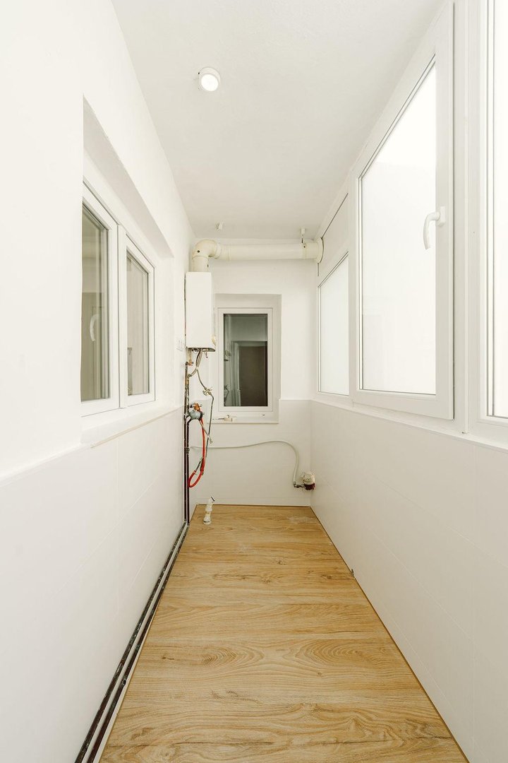 A narrow balcony with wood-look flooring, white walls, large windows, and a wall-mounted water heater unit.