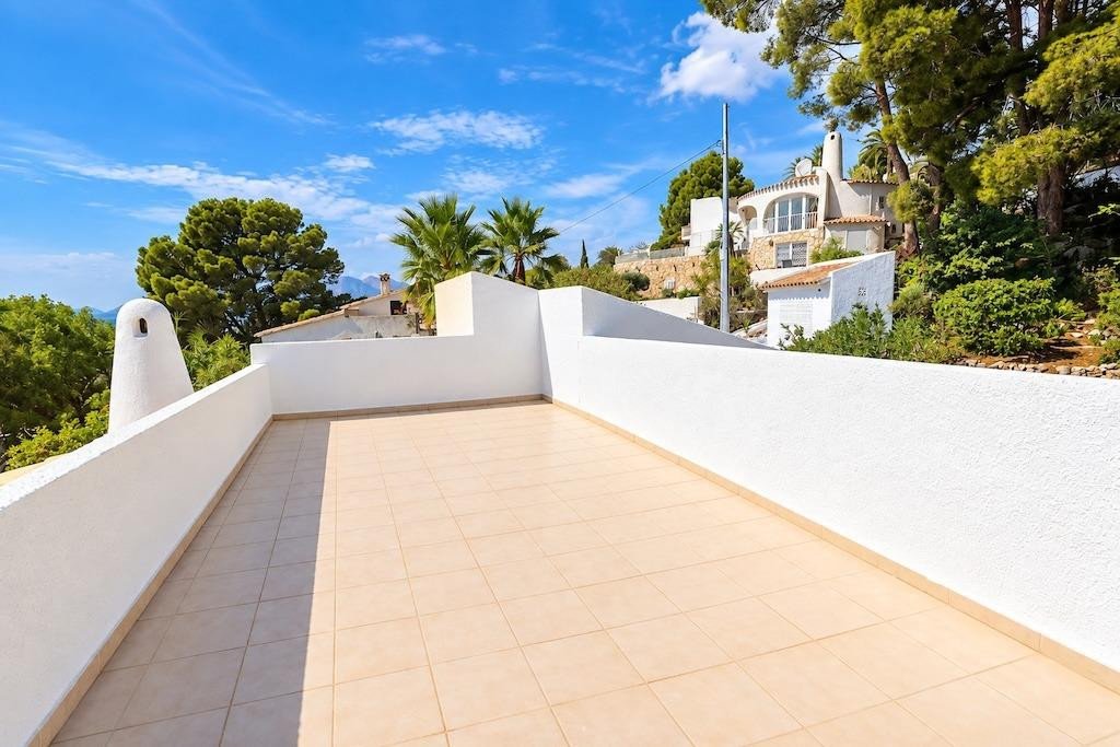 Spacious tiled terrace with white walls, offering a view of trees, houses, and distant hills under a blue sky.