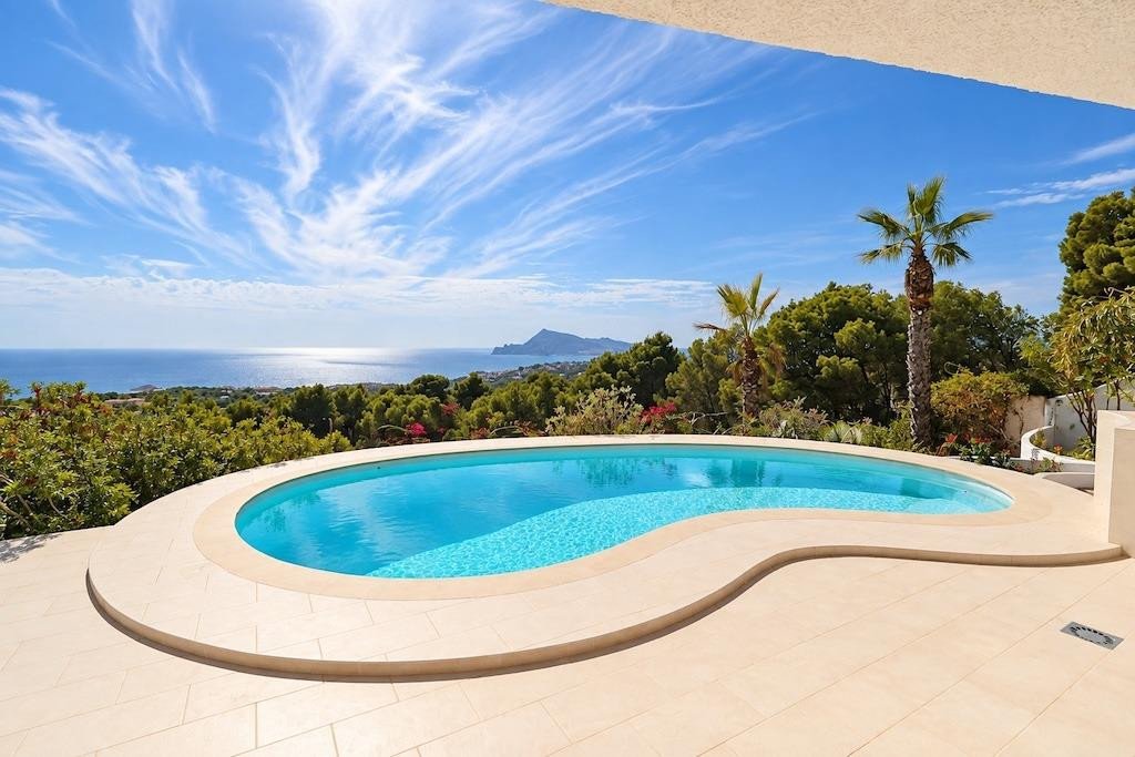 A beautiful swimming pool with a wavy shape, overlooking the sea with distant mountains and lush greenery under a blue sky.