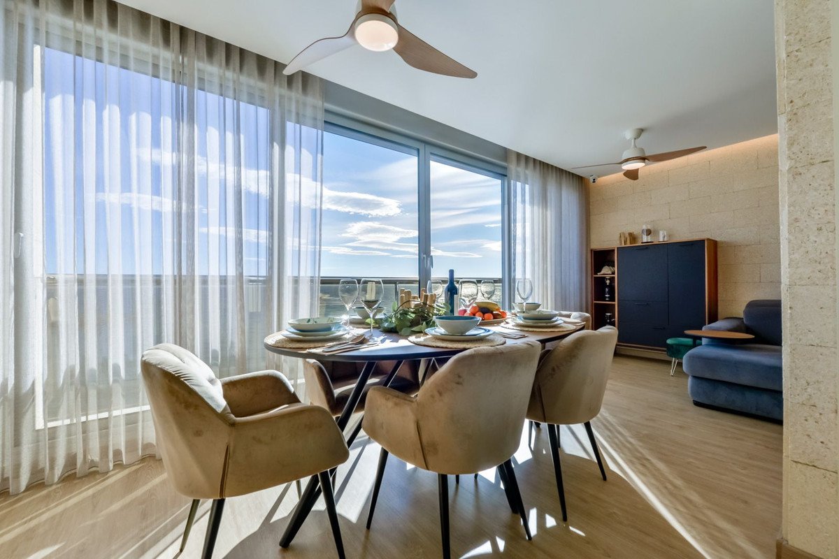 Dining room with table set for four, overlooking the sea from a balcony with floor-to-ceiling windows and sheer curtains.
