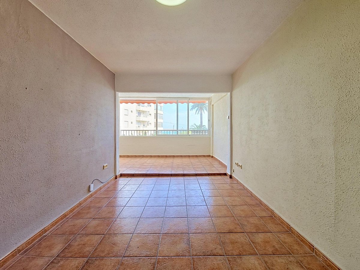 Spacious living area with tiled floor, leading to a sunroom with a balcony overlooking the sea and palm trees.