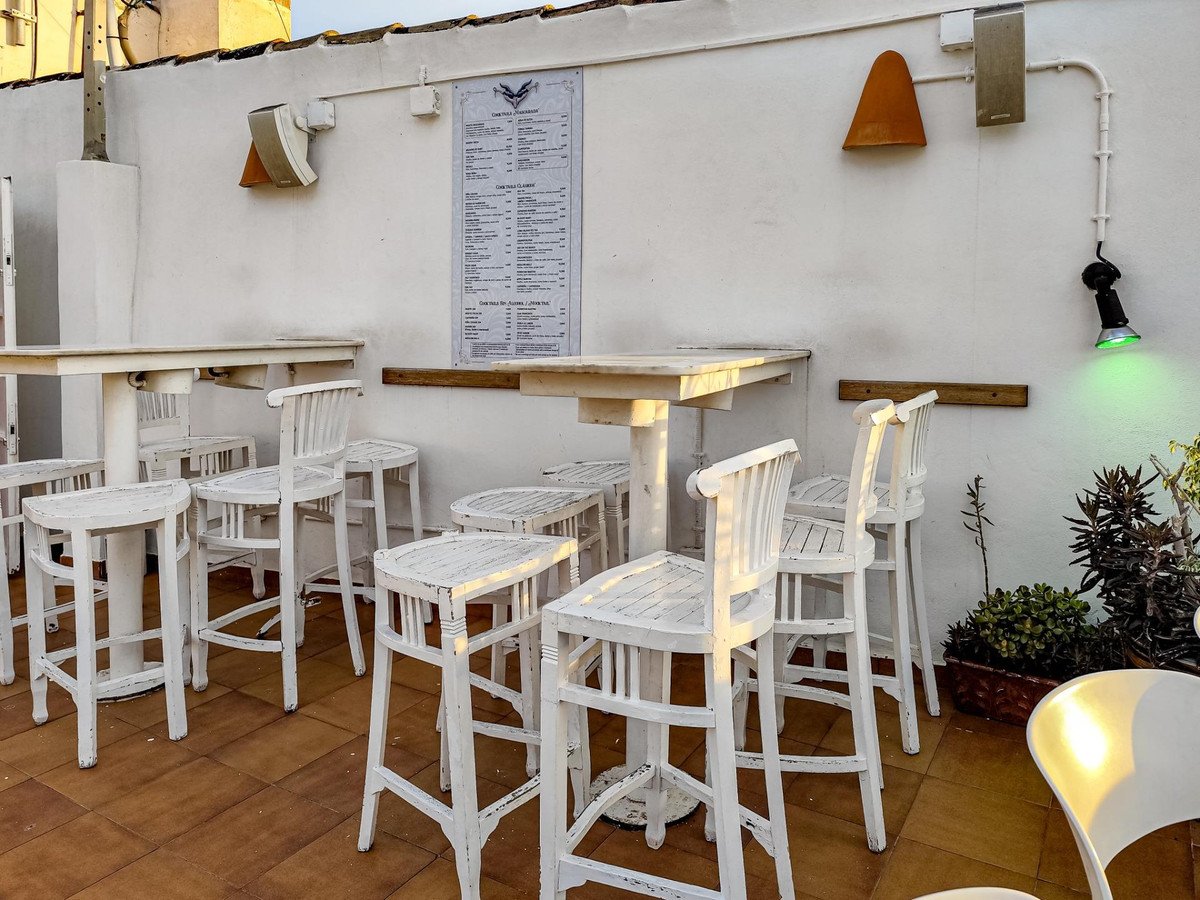 Outdoor rooftop bar seating area with white high chairs and tables, a menu on the wall, and green spotlight.