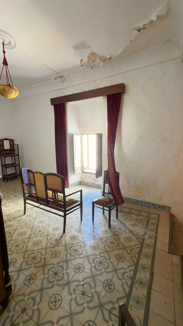 A room with vintage furniture, a bench, chairs, and a dresser by a window with red curtains. Features patterned tiles and peeling ceiling paint.