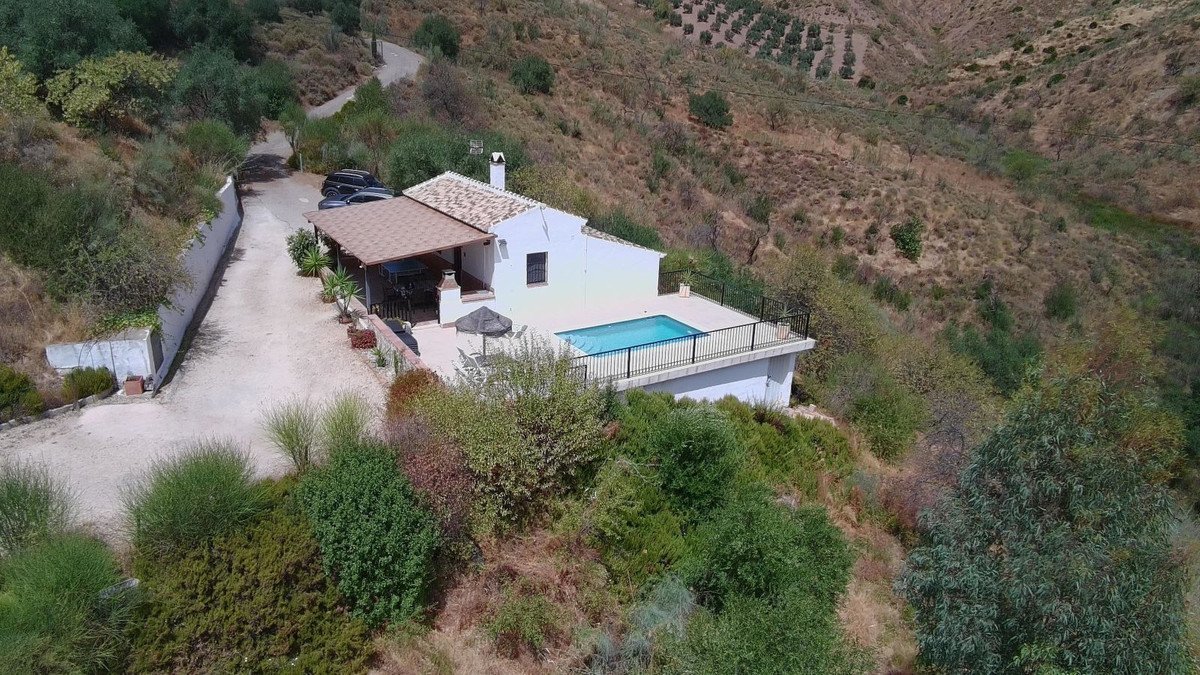 A white villa with a swimming pool and outdoor seating area, set on a hillside with views of olive groves and surrounding nature.