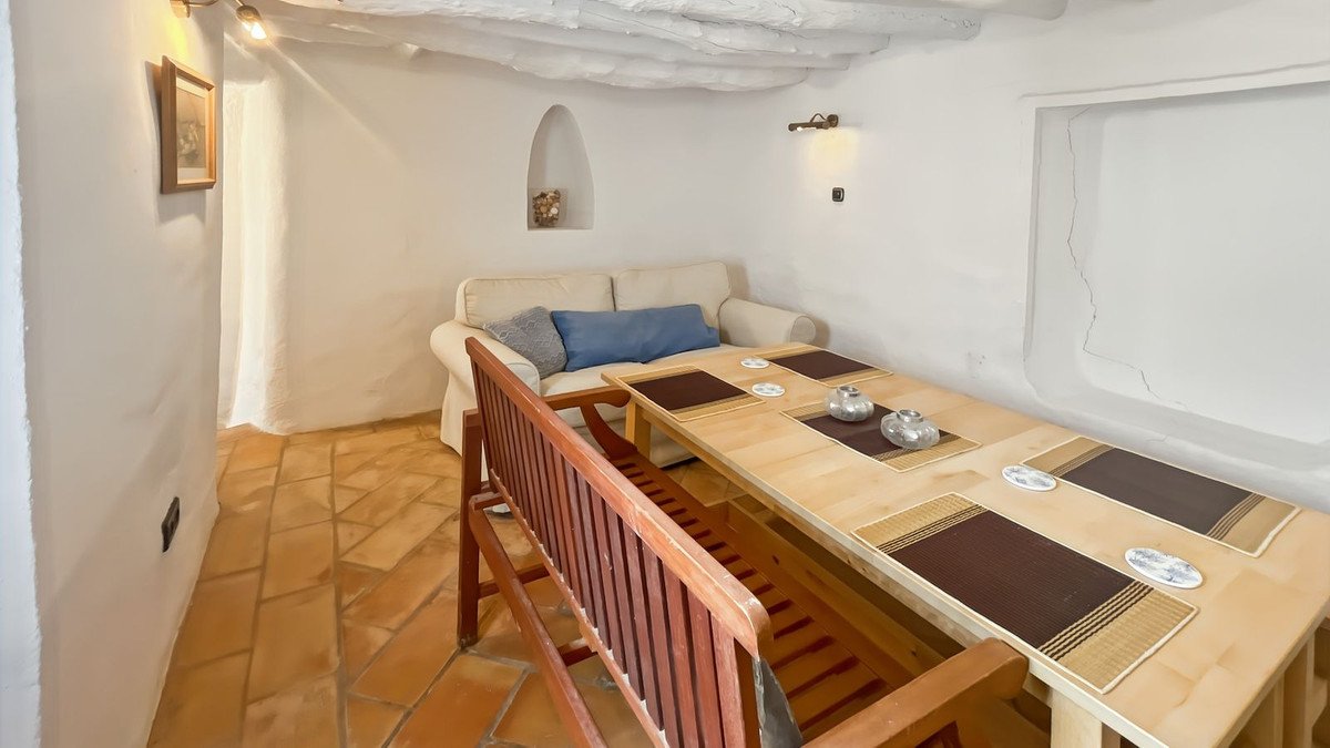 Living and dining area with a sofa, wooden table, benches, and terracotta floor tiles. Rustic, cozy ambiance.