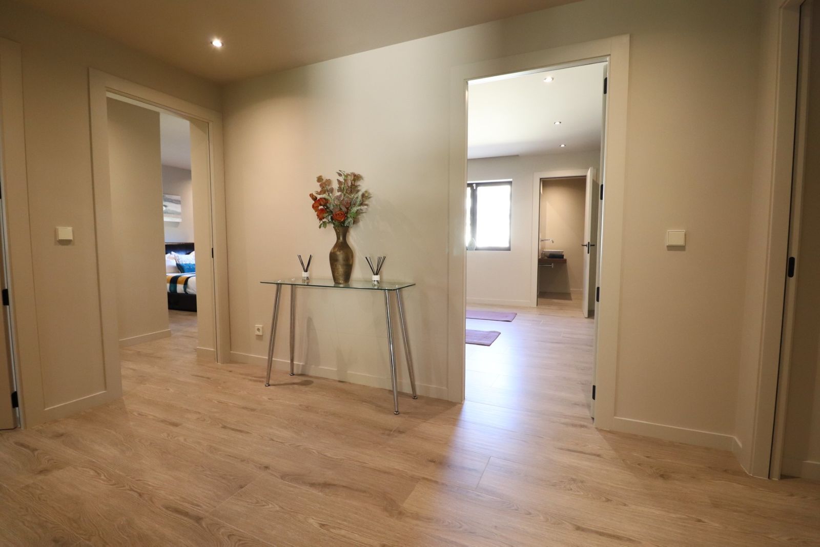 Moderne hal met glazen tafel, bloemenvaas en doorkijk naar slaapkamer en badkamer.