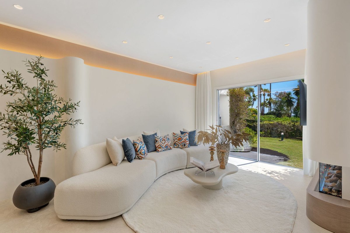 Modern living room with curved sofa, plush rug, and a sliding glass door leading to a lush garden view with palm trees.