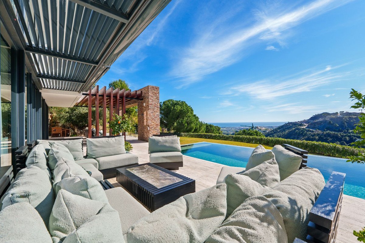 Modern patio with outdoor seating and infinity pool overlooking the sea and hills under a clear blue sky.