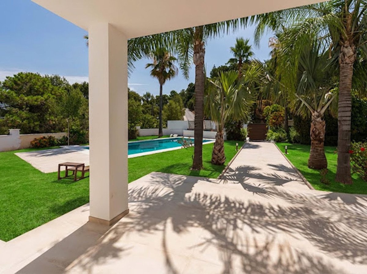 View from a covered patio looking out to a swimming pool, manicured lawn, and lush palm trees under a sunny sky.