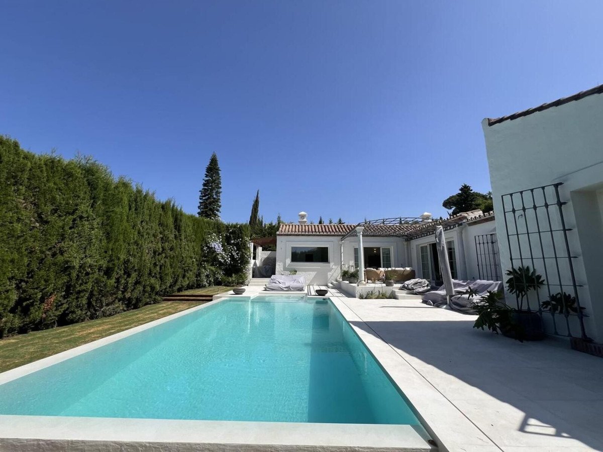 A spacious villa with a bright blue swimming pool and outdoor seating area, surrounded by lush greenery and a clear blue sky.