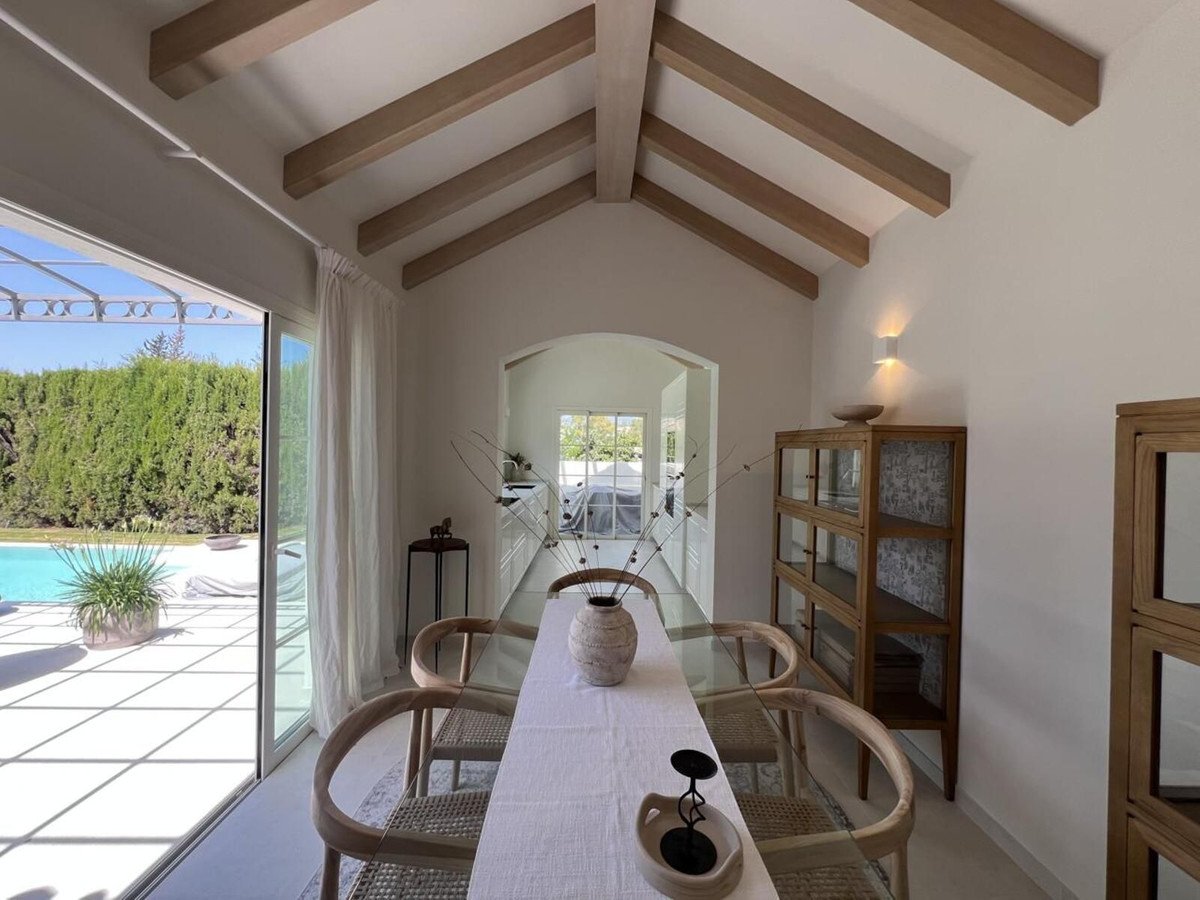 Modern dining room with glass table, wooden chairs, and a view of a swimming pool and garden through large sliding doors.