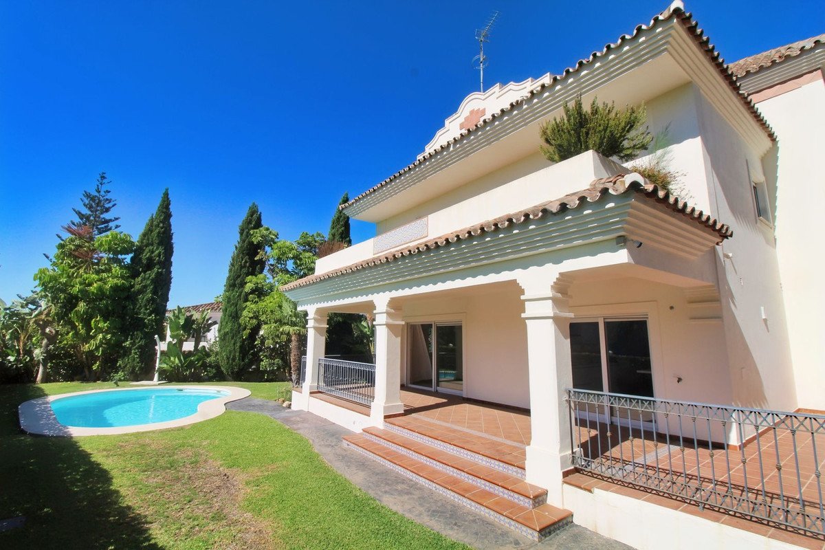 A modern villa with a covered patio, a kidney-shaped swimming pool, and a lush green garden under a clear blue sky.