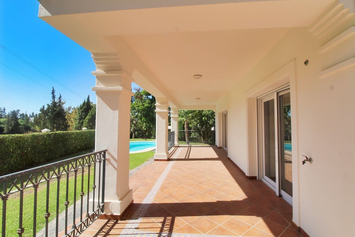 Covered terrace with a view of a swimming pool and garden, featuring white columns and tiled flooring.