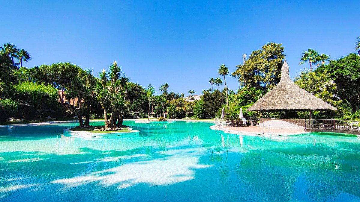Luxurious swimming pool with a thatched-roof bar and tropical greenery under a clear blue sky.