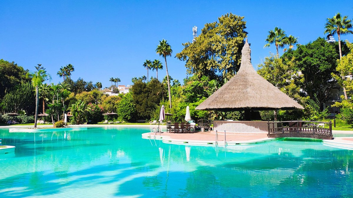 A large, turquoise swimming pool with a thatched roof bar and seating area, surrounded by lush tropical greenery and palm trees.
