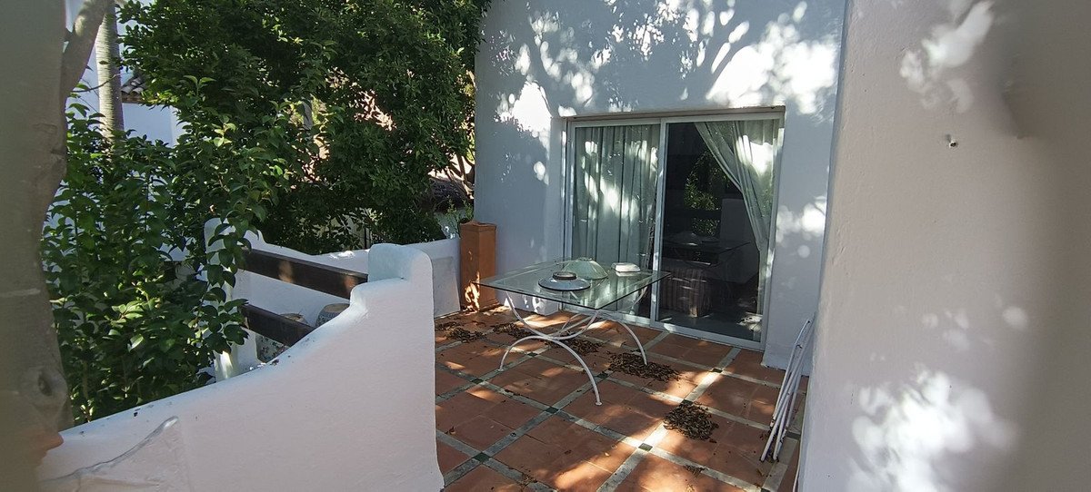 A sunny terrace with a glass table, chairs inside, and lush green trees providing shade. A sliding glass door leads indoors.