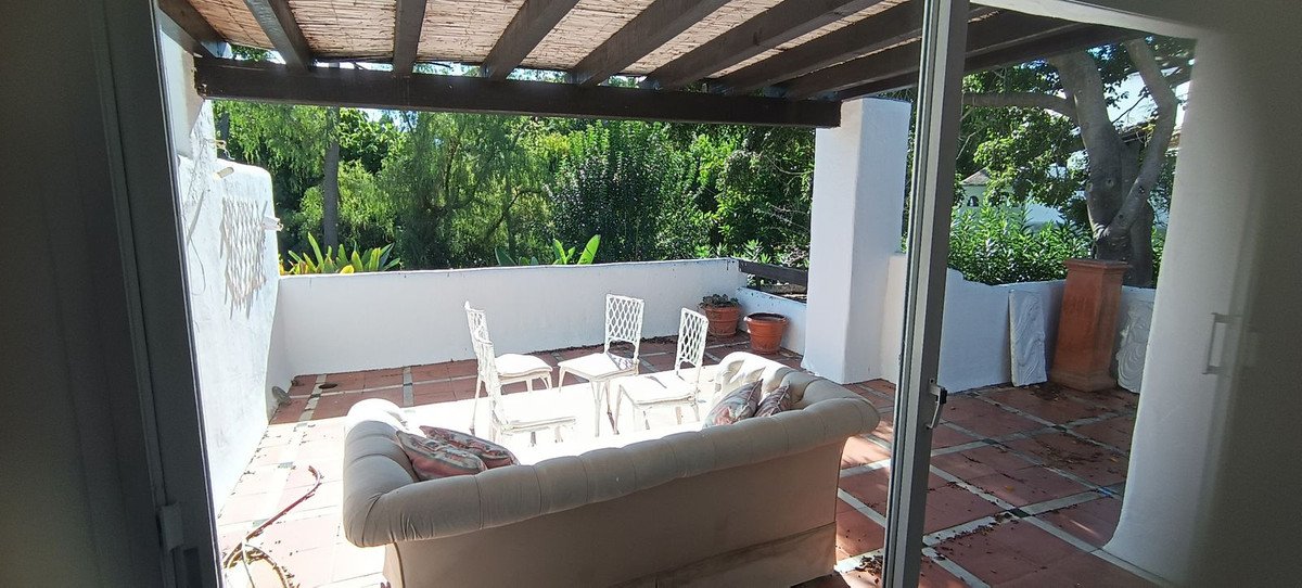 Covered terrace with sofa, chairs, and potted plants, overlooking lush greenery and trees.