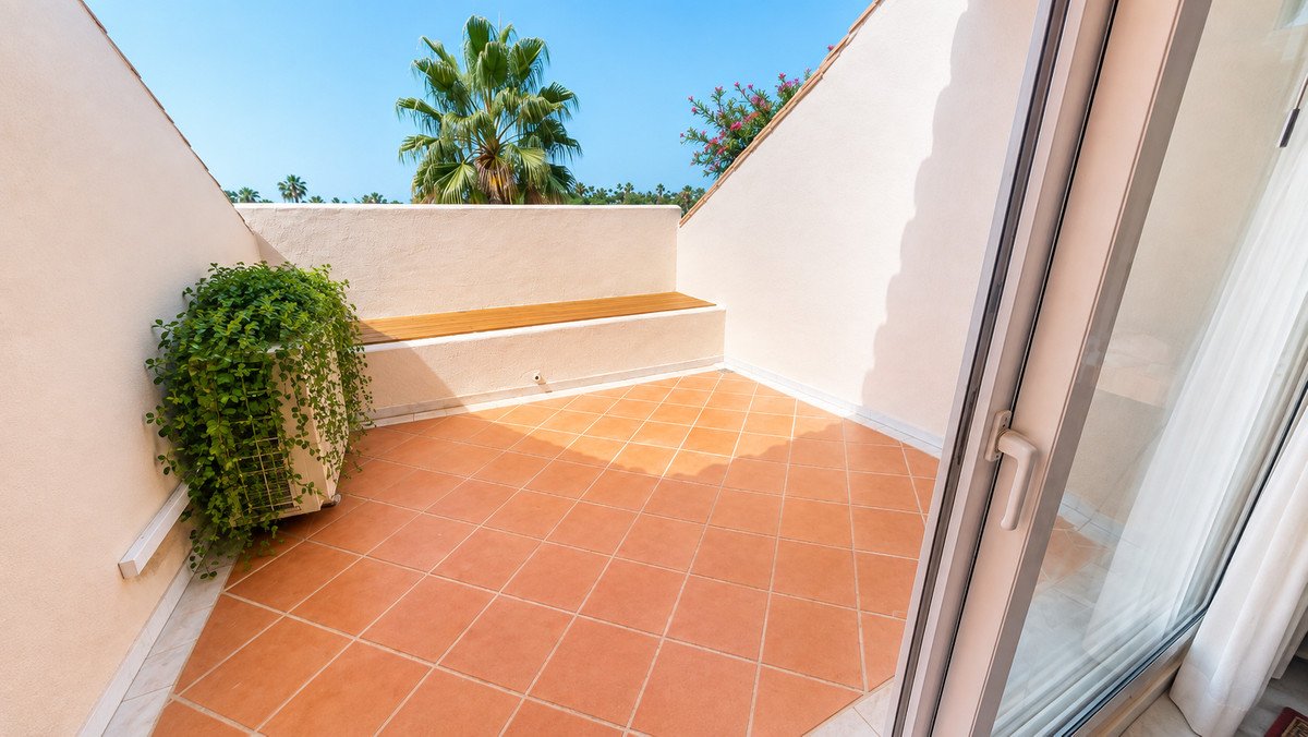 An empty tiled terrace with a built-in bench, potted plants, and palm trees visible in the background.