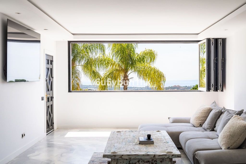 Modern living room with large window view of palm trees and ocean, grey sofa, marble floor.