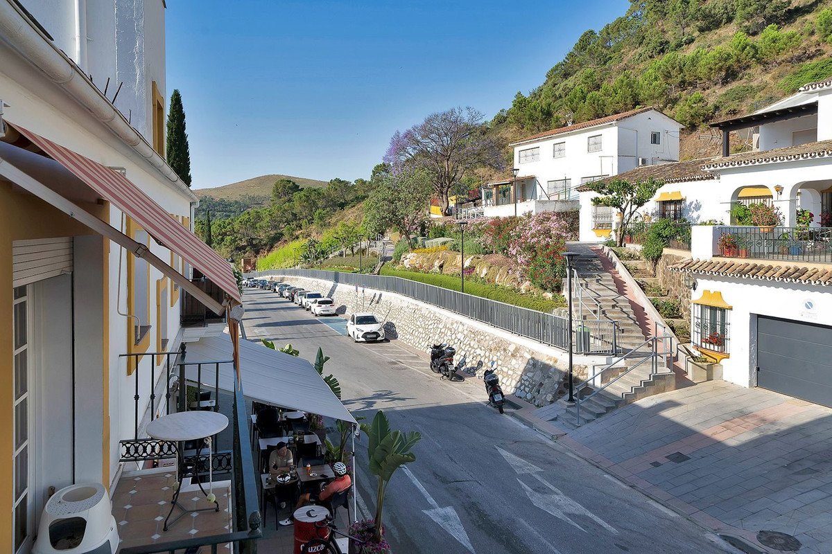 Apartment with balcony overlooking a sunny street, white buildings, and green hills.