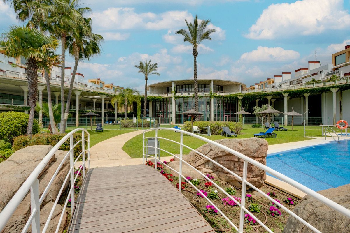 Wooden bridge leads to pool area with palm trees, lounge chairs, and modern building under blue sky.