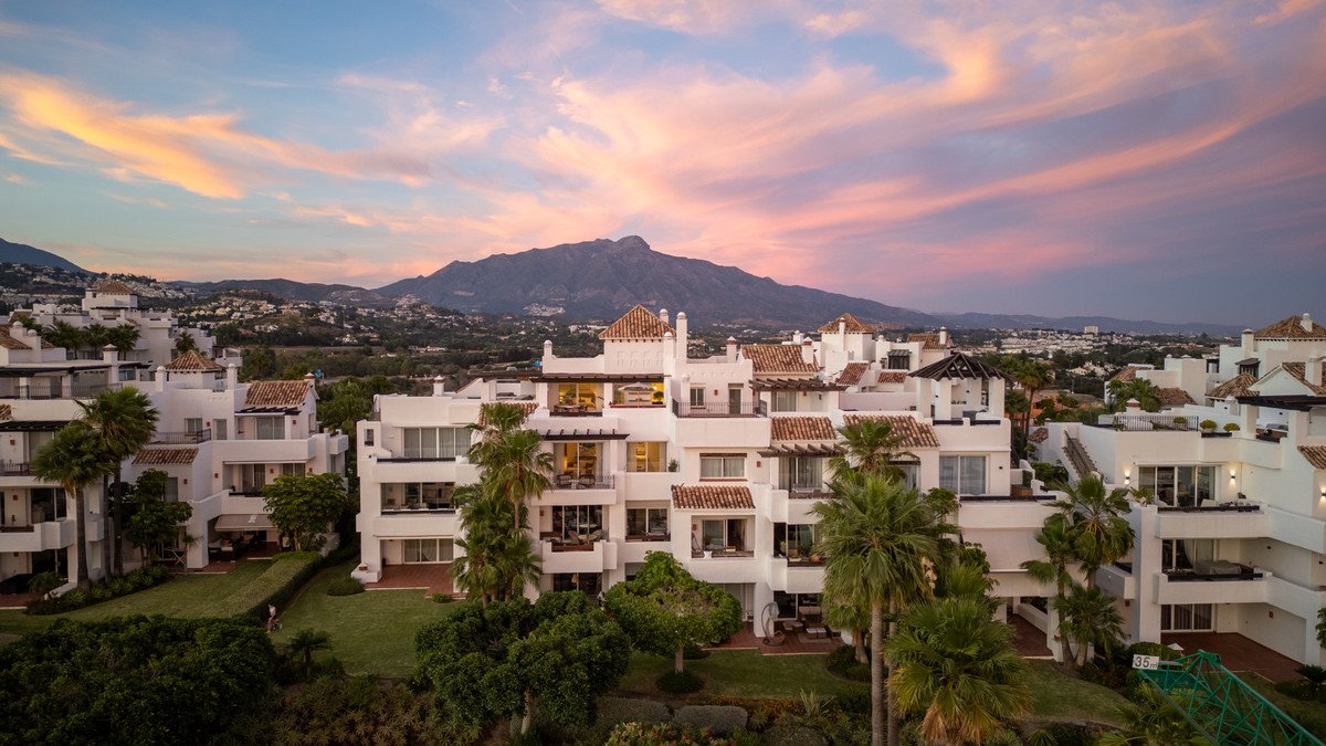 Modern white villa with terracotta roofs, palm trees, and mountain views at sunset.