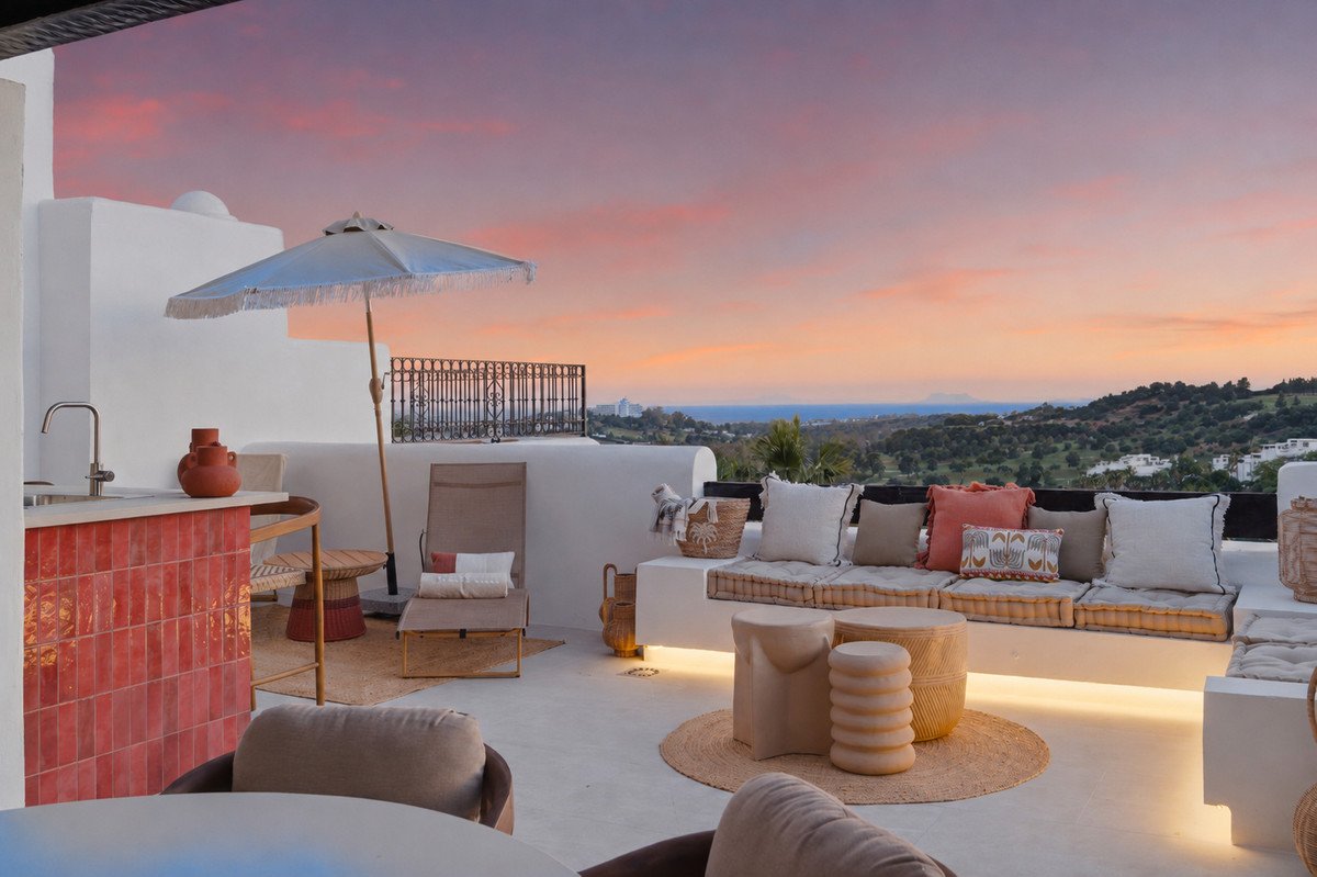 Rooftop terrace with lounge seating, red tile bar, and sunset view over hills and sea.
