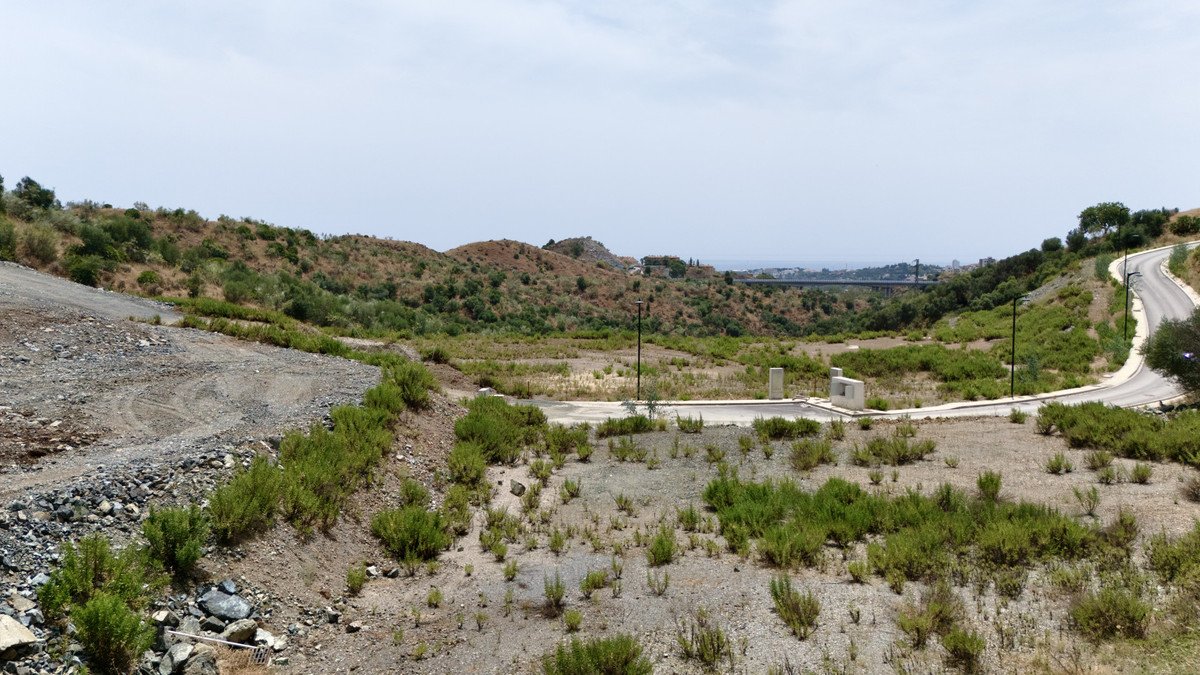 A vacant lot with gravel, shrubs, and a winding road, overlooking hills and a distant bridge under a cloudy sky.